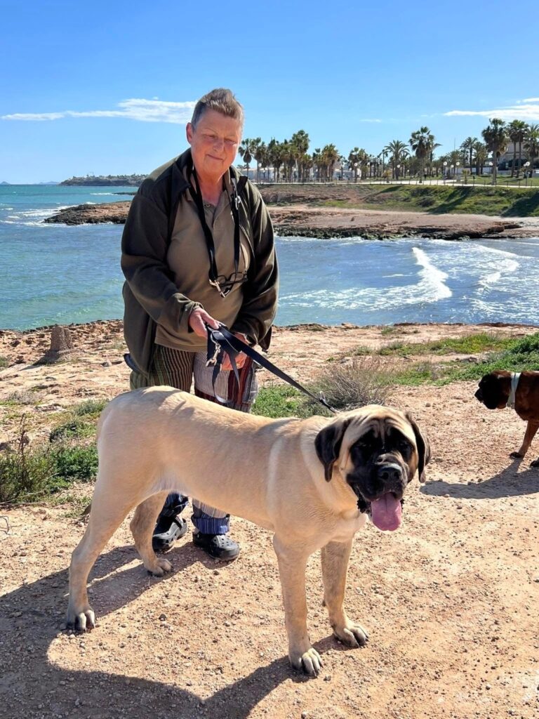 Sybilla steht mit der fawnfarbenen English-Mastiff-Hündin Greta an einer sonnigen Meeresküste.