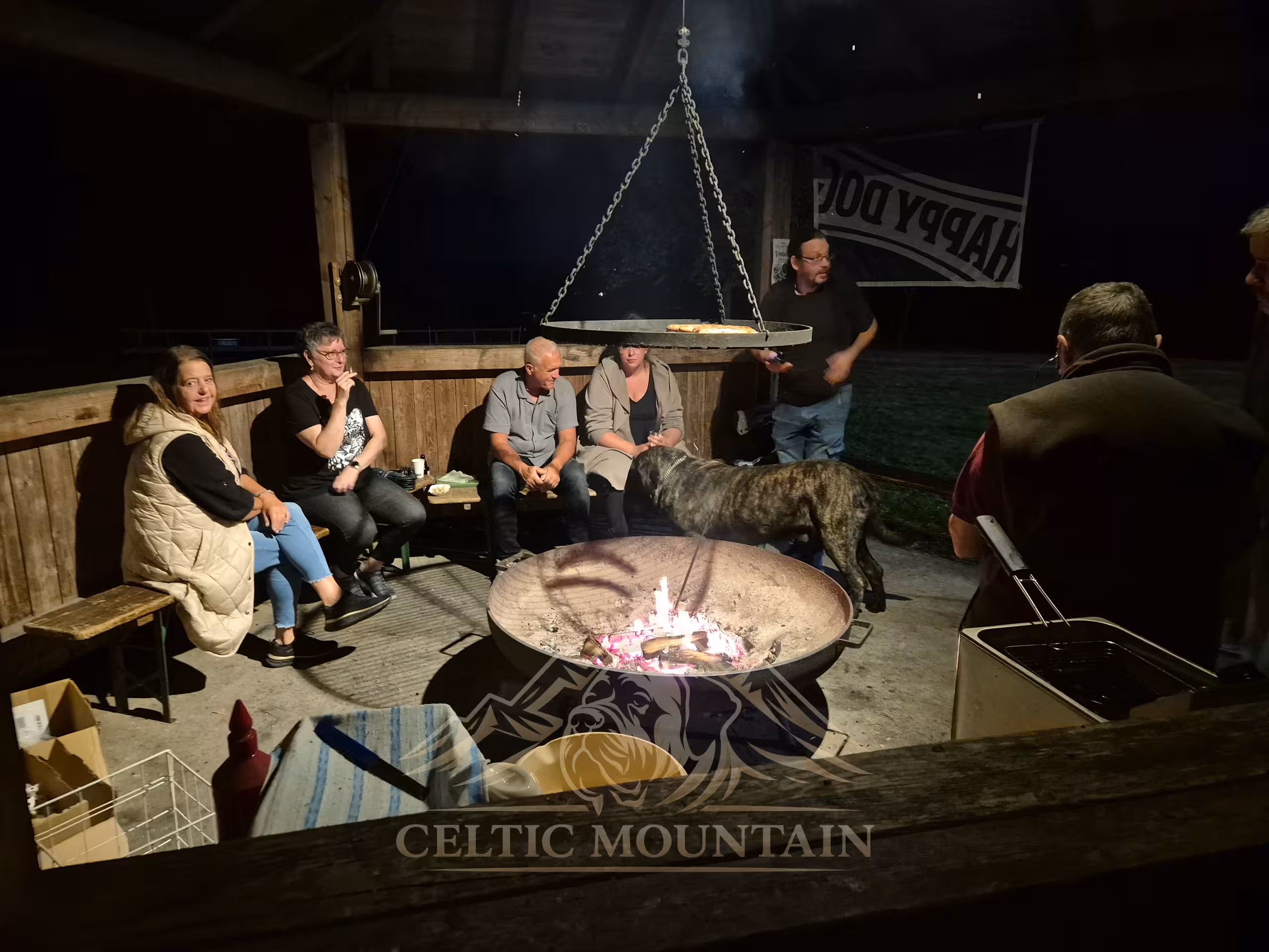 Guests sit around a burning fire bowl inside the wooden pavilion while a brindle Mastiff stands beside them.