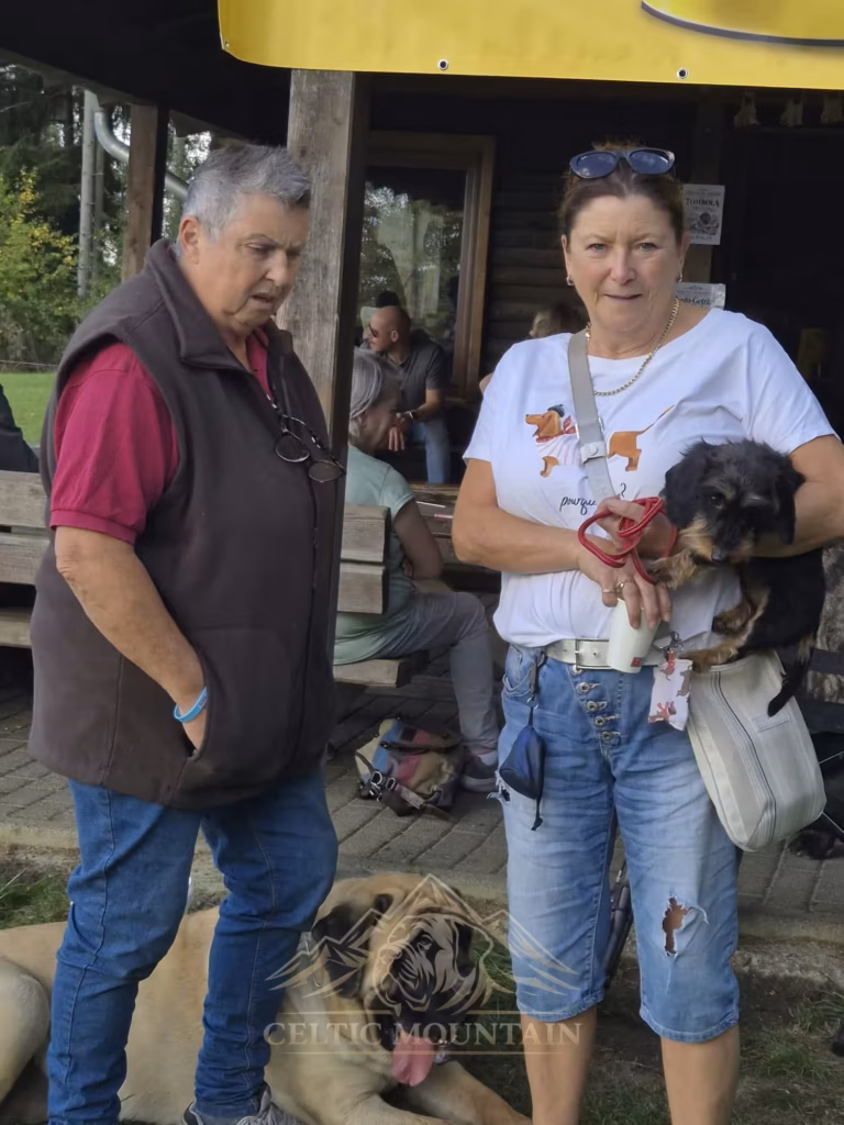 Zwei Gäste stehen vor der Grillhütte; ein kleiner dunkler Hund wird getragen, ein Mastiff liegt zu ihren Füßen.
