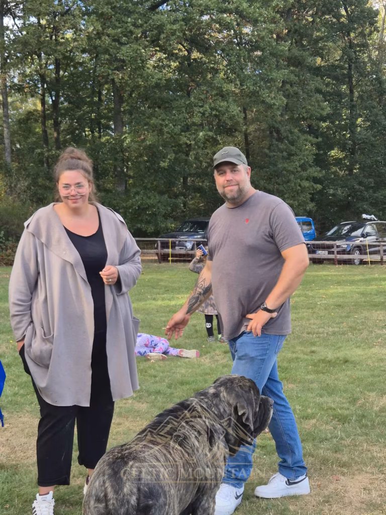 Zwei Gäste lächeln auf der Wiese zur Kamera; ein gestromter Mastiff steht vor ihnen.