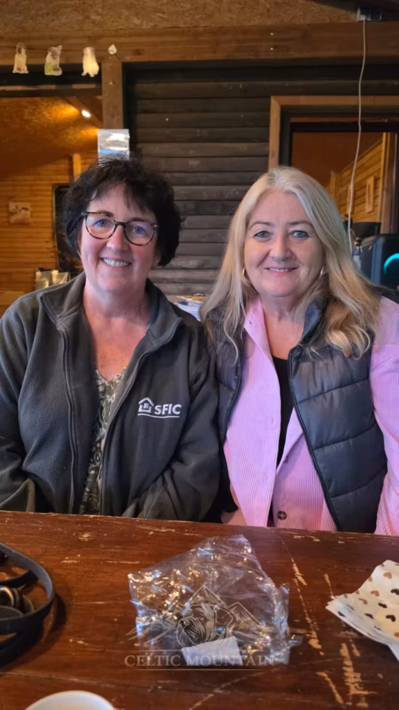 Zwei Gäste lächeln nebeneinander an einem Tisch in der Holzgrillhütte zur Kamera.