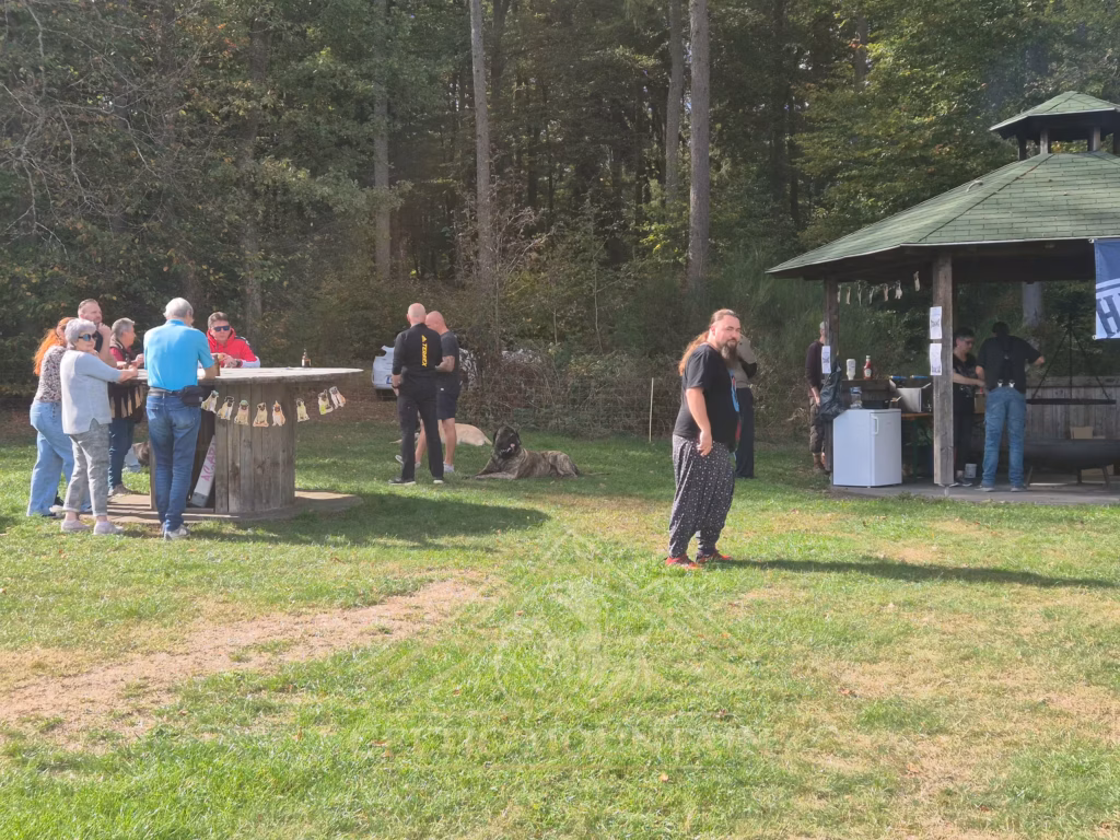 Mehrere Gäste stehen auf der Wiese zwischen rundem Holztisch, Pavillon und Waldrand.