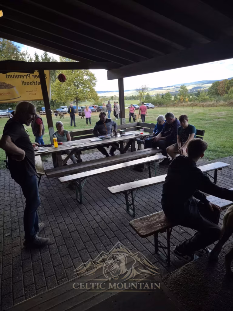 Mehrere Gäste sitzen an langen Tischen im Schatten der Grillhütte; draußen liegen Hunde auf der Wiese.