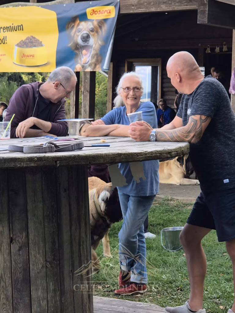Drei Gäste stehen an einem runden Holztisch vor der Grillhütte und sprechen miteinander.